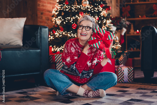 Full length portrait of cheerful minded lady sit carpet floor hold desirable christmas giftbox imagine evergreen tree lights indoors
