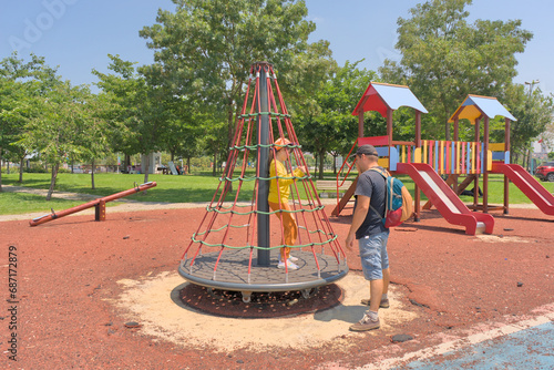 A child on a children's carousel is spun by his father on a playground in a park on a clear sunny day.
