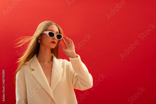 Fashionable confident woman wearing trendy sunglasses, white boucle coat, posing on red background. Close up studio portrait. Copy, empty space for text