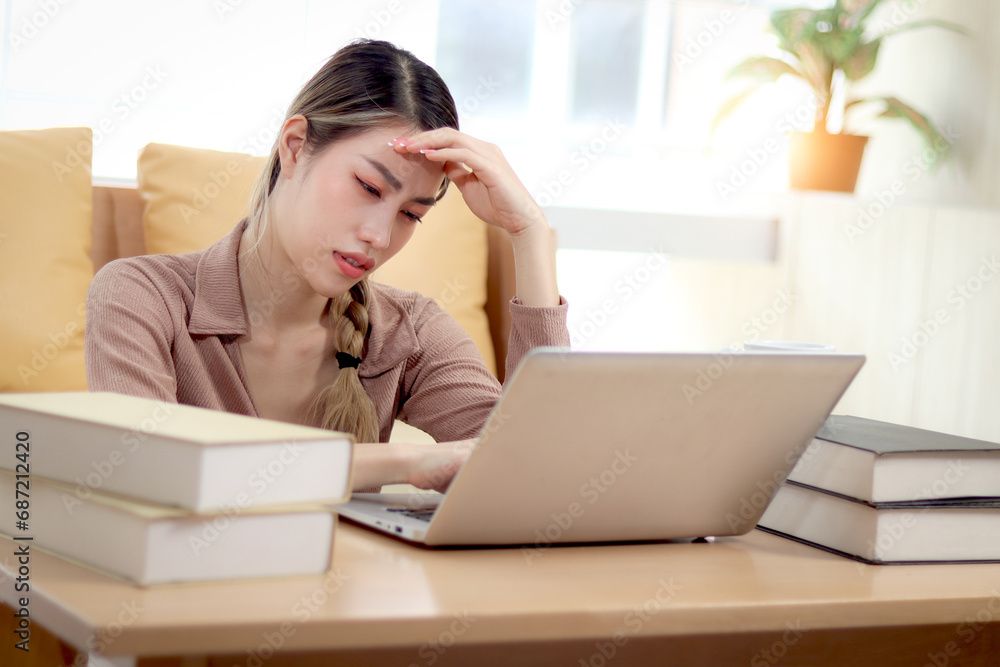 Serious tired beautiful Asian woman putting head in hand during using laptop computer. Thoughtful confused young female student feeling stressed, having headaches while studying of a test at home. Serious tired beautiful Asian woman putting head in hand during using laptop computer. Thoughtful confused young female student feeling stressed, having headaches while studying of a test at home.