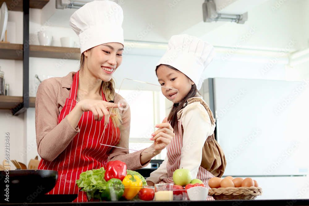 Happy Asian mother and daughter girl wear apron chef hat at kitchen, mom touch transparent device tablet for searching tasty meal recipes on internet, cute kid chef and family cooking food at home. Happy Asian mother and daughter girl wear apron chef hat at kitchen, mom touch transparent device tablet for searching tasty meal recipes on internet, cute kid chef and family cooking food at home.