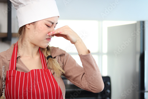 Unhappy irritated glamour Asian beautiful woman with apron chef and hat putting fingers to cover her nose at kitchen. Young female chef cannot tolerate bad smell and pungent while cooking food.