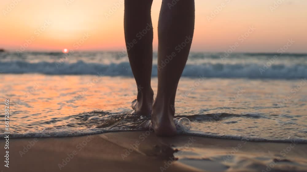 A girl walks barefoot along the beach at sunset, leaving footprints on the sand and reflections in the water. close-up of female legs on the beach. calmness and tranquility. tourist on summer vacation