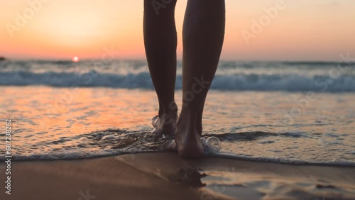 A girl walks barefoot along the beach at sunset, leaving footprints on the sand and reflections in the water. close-up of female legs on the beach. calmness and tranquility. tourist on summer vacation photo