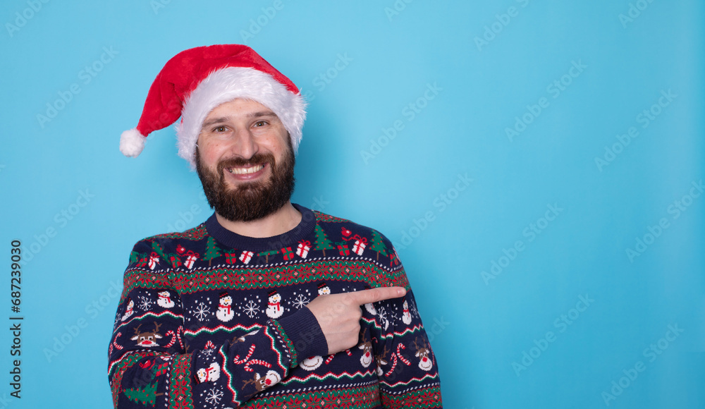 Handsome caucasian bearded man in red Christmas cap pointing to free space and looking at camera isolated over blue background. Christmas sale. Handsome caucasian bearded man in red Christmas cap pointing to free space and looking at camera isolated over blue background. Christmas sale.