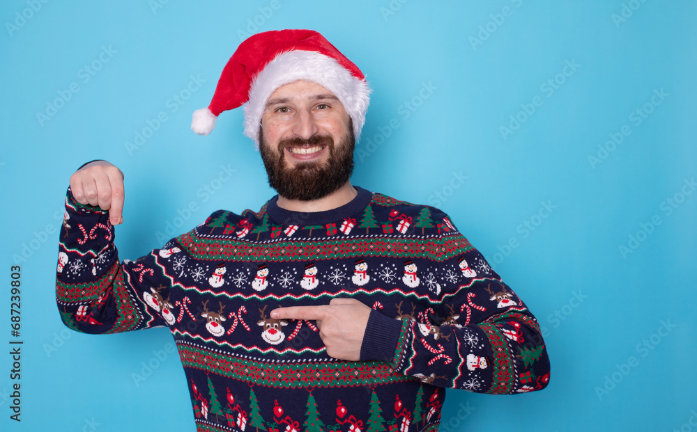 Handsome caucasian bearded man in red Christmas cap pointing to free space and looking at camera isolated over blue background. Christmas sale.
