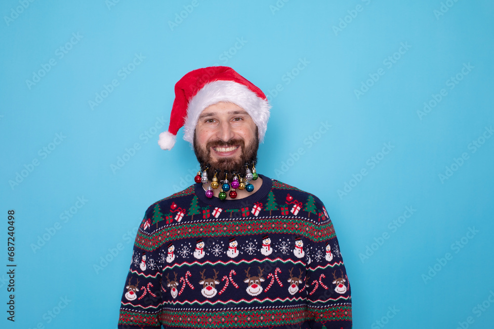 Handsome bearded caucasian guy in sweater and Santa hats stands on blue background in studio and teeth smiling. Portrait european guy with Christmas mood. Holiday banner. New Year. Handsome bearded caucasian guy in sweater and Santa hats stands on blue background in studio and teeth smiling. Portrait european guy with Christmas mood. Holiday banner. New Year.