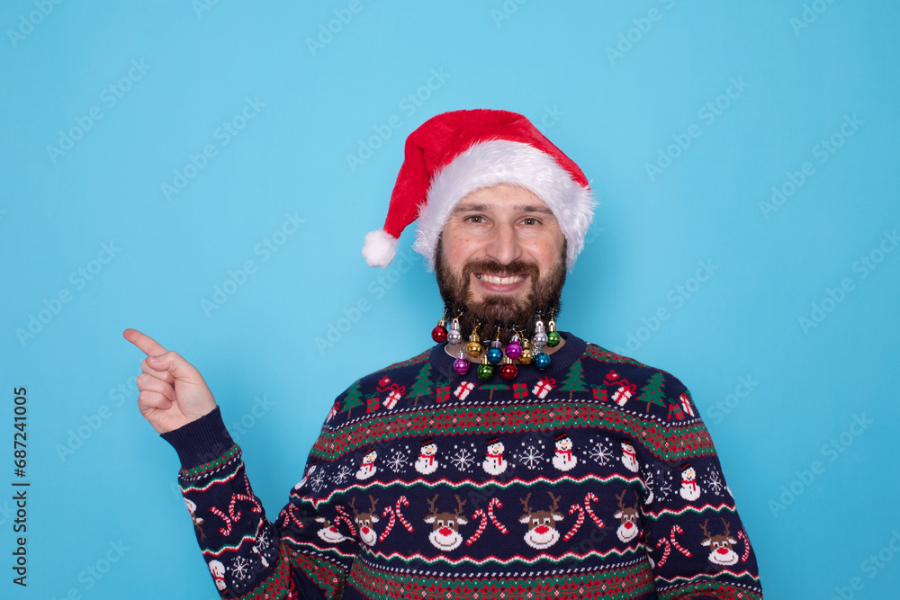 Handsome caucasian bearded man in red Christmas cap pointing to free space and looking at camera isolated over blue background. Christmas sale. Handsome caucasian bearded man in red Christmas cap pointing to free space and looking at camera isolated over blue background. Christmas sale.