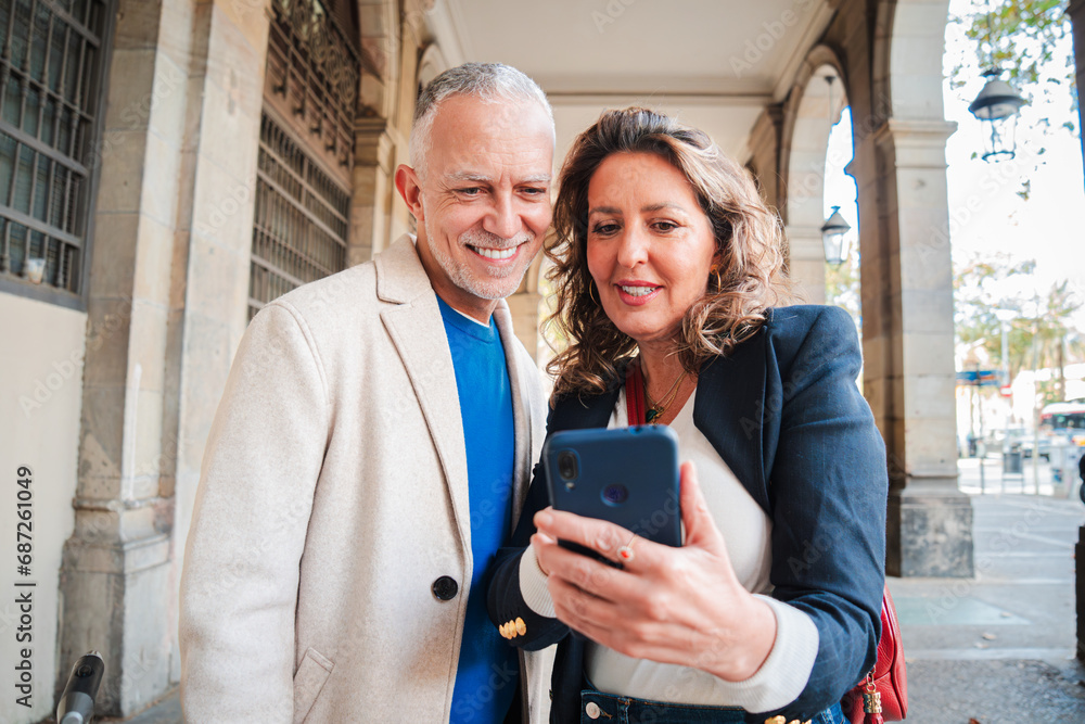 Fototapeta premium Mature tourists couple using a smartphone and watching funny pictures on their weekend trip and enjoying together. Middle aged woman and her husband searching hotel on a cell phone standing outside