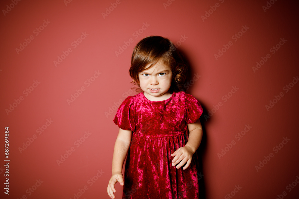 Pretty caucasian brunette toddler little girl in red velour dress dissatisfied and angry at the red background. Christmas vibe Pretty caucasian brunette toddler little girl in red velour dress dissatisfied and angry at the red background. Christmas vibe