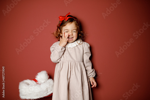 Adorable caucasian brunette toddler little girl in beige dress and red ribbon smiling widely with the finger in noise at the red background. Christmas vibe 