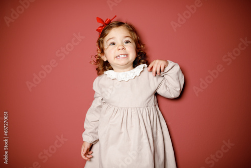 Adorable caucasian brunette toddler little girl in beige dress and red ribbon smiling widely at the red background. Christmas vibe 