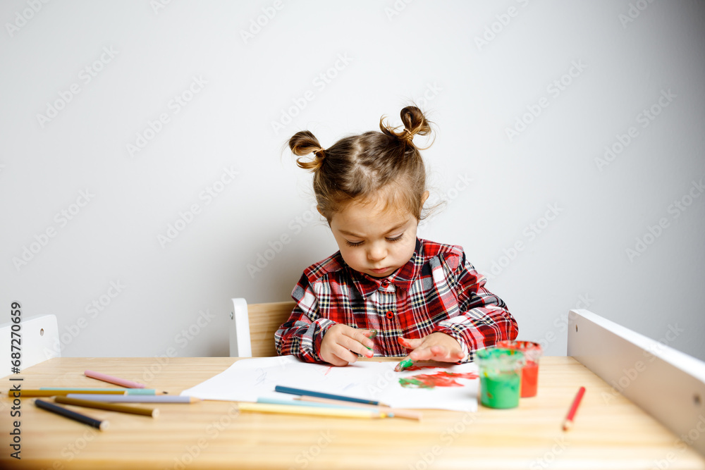 Portrait of cute toddler girl with two bunches in checkered dress sitting on the baby wooden table and chair and drawing with red finger paints with messy hands, Montessori concept fine motricity Portrait of cute toddler girl with two bunches in checkered dress sitting on the baby wooden table and chair and drawing with red finger paints with messy hands, Montessori concept fine motricity