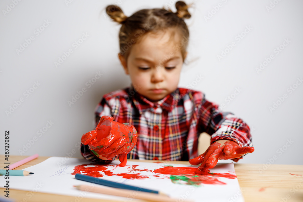 Portrait of cute toddler girl with two bunches in checkered dress sitting on the baby wooden table and chair and drawing with red finger paints with messy hands, Montessori concept fine motricity Portrait of cute toddler girl with two bunches in checkered dress sitting on the baby wooden table and chair and drawing with red finger paints with messy hands, Montessori concept fine motricity