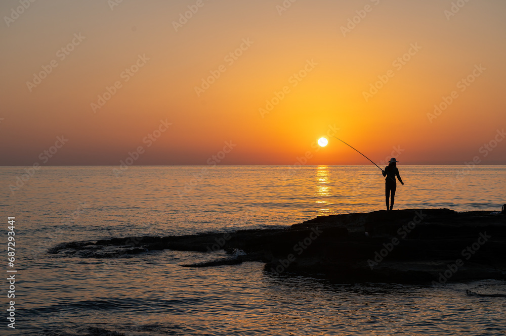 Silhouette of a girl fishing with a fishing rod in the sea at sunset.