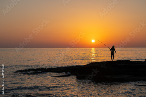 Silhouette of a girl fishing with a fishing rod in the sea at sunset.