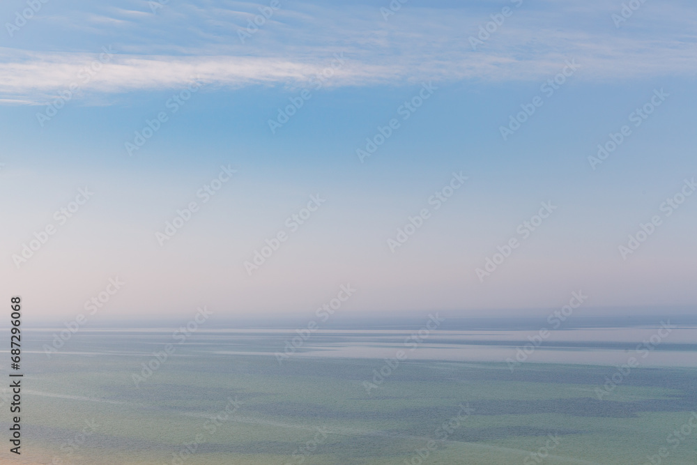 Blue sky with haze and blurry glowing pink clouds above the sea. Shallow coastal sea water with azure patterns. Ripples in the water