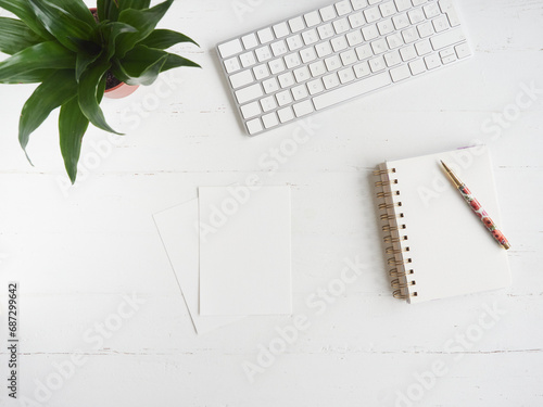 notebook, blank sheets of paper and pen on white desk table beside a computer keyboard and a green plant as decoration. Top view with copy space, flat lay.