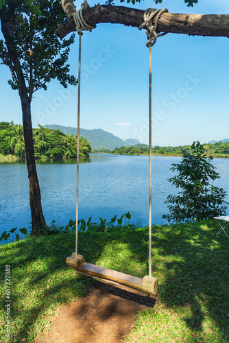 Wooden rope swing wood seat hanging from tree branch with lake view and blue sky background  Kanchanaburi  Thailand.