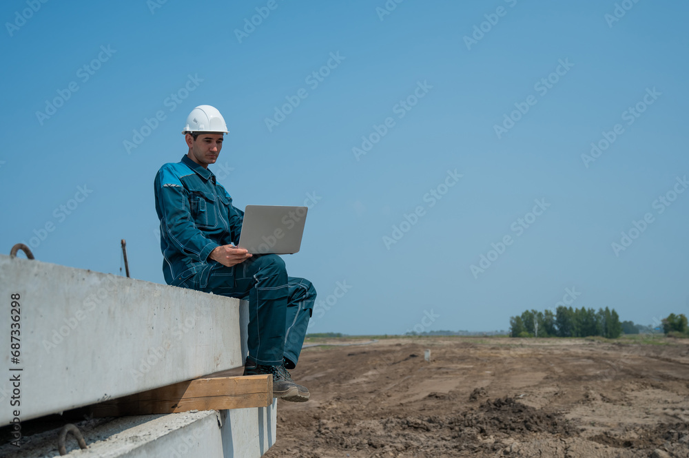 Caucasian male builder in hardhat sits on floor slabs and uses laptop at construction site. Caucasian male builder in hardhat sits on floor slabs and uses laptop at construction site.