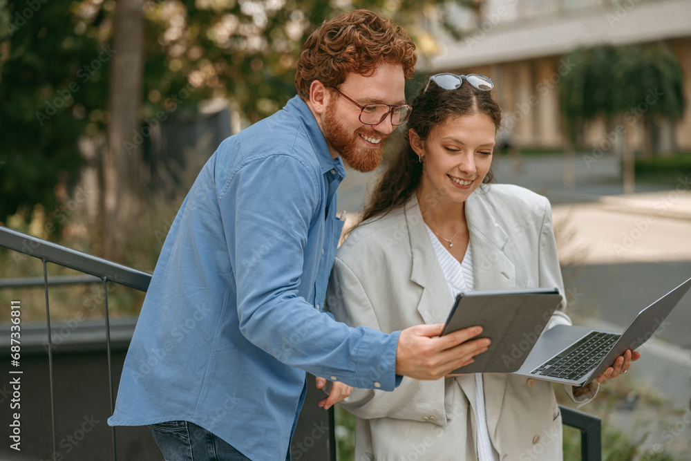 Smiling business colleagues discuss biz issue while use laptop standing on building background