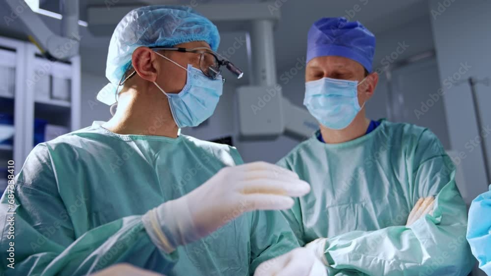 Adult surgeon wearing device glasses talks to his colleagues standing beside and listening to him carefully. Low angle view on the male doctors in surgery room. Adult surgeon wearing device glasses talks to his colleagues standing beside and listening to him carefully. Low angle view on the male doctors in surgery room.