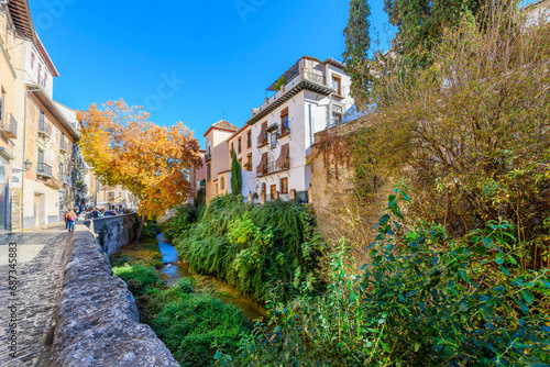 Pedestrians walk the narrow, historic Carrera del Darro street past shops, medieval buildings and the Darro river in the picturesque center of the Andalusian city of Granada, Spain at autumn. 