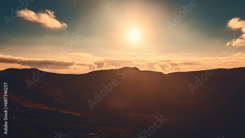 Sunrise cloudscape sky over mountain range silhouette. Dramatic clouds flow in dark sky, sun glow rise over mountain peak with golden light beams. Beautiful morning summer landscape.