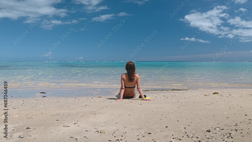 Woman relax sitting on sand beach enjoy blue sky and crystal sea water. Caucasian girl in bikini with snorkeling mask, back view. Outdoor lifestyle travel on summer holiday vacation.