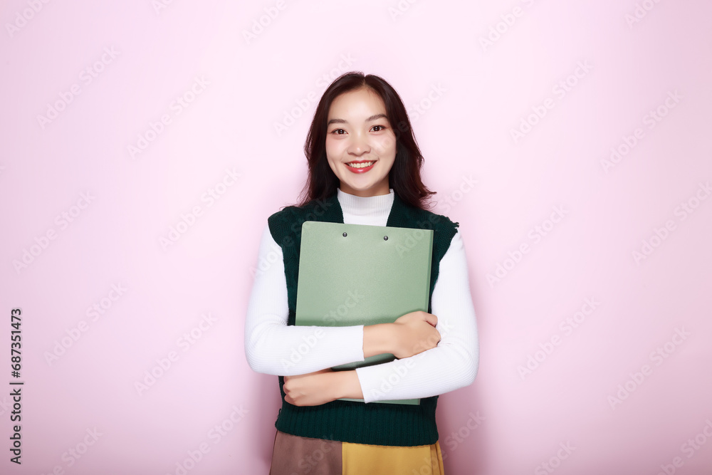 Asian woman poses holding a file folder while standing in front of a light pink background. Asian woman poses holding a file folder while standing in front of a light pink background.