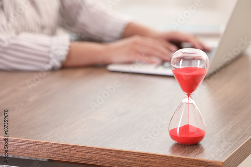 Hourglass with red flowing sand on table. Woman using laptop indoors, closeup