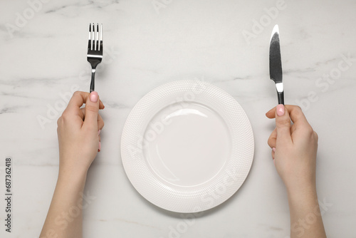 Woman holding fork and knife near empty plate at light marble table, top view