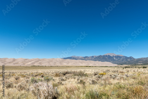 Desert landscape of shrub grassland, sand dunes, and mountains under a clear blue sky; taken outside of Great Sand Dunes National Park in Colorado.