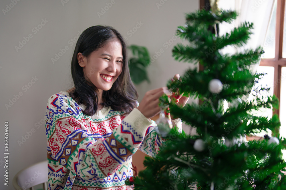 Young asian woman in sweater hanging and decorating candy cane with toys ornaments on christmas tree while preparing christmas tree to celebrate for xmas festive holiday and winter seasons at house Young asian woman in sweater hanging and decorating candy cane with toys ornaments on christmas tree while preparing christmas tree to celebrate for xmas festive holiday and winter seasons at house