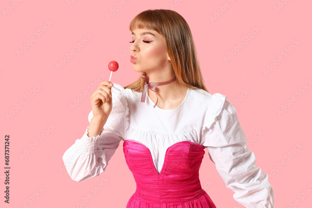 Young woman in doll clothes with lollipop blowing kiss on pink background Young woman in doll clothes with lollipop blowing kiss on pink background