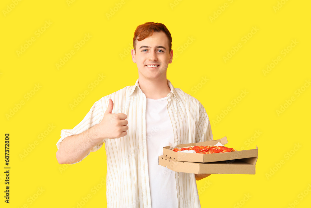 Young man holding cardboard box with tasty pizza and showing thumb-up on yellow background Young man holding cardboard box with tasty pizza and showing thumb-up on yellow background