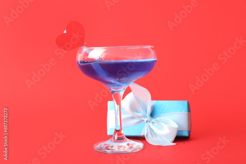 Glass of tasty cocktail with gift on red background. Valentine's Day celebration