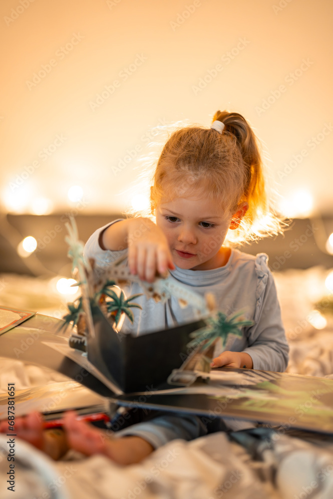 Fototapeta premium Little girl reading pop up book in home bed in Christmas environment with lights background