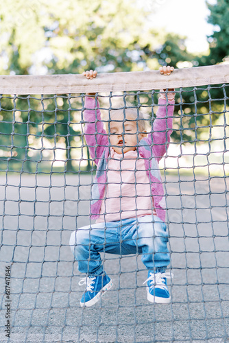 Little girl hangs on the edge of a tennis net with her legs folded
