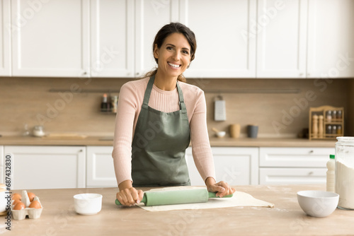Happy beautiful female baker woman in apron home portrait. Cheerful young woman baking pastry in kitchen, looking at camera with toothy smile, enjoying domestic work, dessert preparation