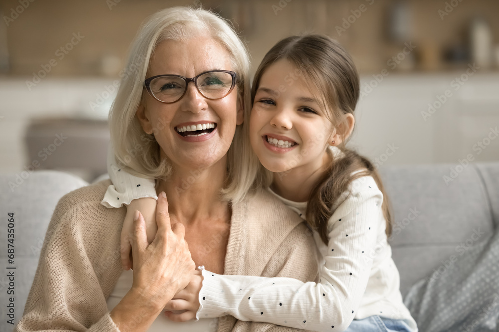 Happy pretty tween granddaughter girl hugging loving grandma from behind, looking at camera, smiling, laughing, enjoying leisure with granny, posing for family portrait Happy pretty tween granddaughter girl hugging loving grandma from behind, looking at camera, smiling, laughing, enjoying leisure with granny, posing for family portrait