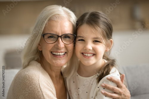 Cheerful beautiful senior grandmother and happy little granddaughter home family portrait. Positive loving granny hugging cute kid with tenderness, affection, looking at camera with toothy smile