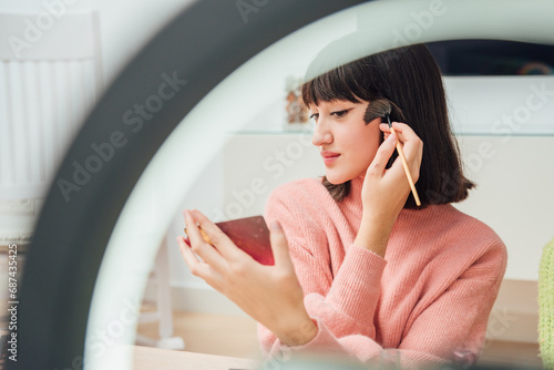 Smiling young ethnic woman sitting with mirror in hand and applying facial brush