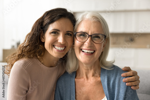 Cheerful beautiful adult daughter woman hugging happy blonde senior mom, holding shoulder with support, care, looking at camera with perfect toothy smile, laughing. Elderly mother headshot portrait