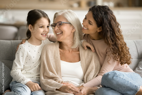 Cheerful grandma, mom and grandkid girl resting on home couch, talking, laughing, having fun, enjoying family leisure, warm close relationship, happy childhood, retirement