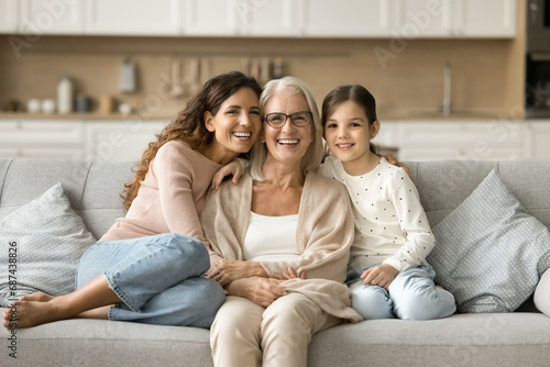 Happy grandma, mom and granddaughter kid sitting close on home sofa. Young mother and girl hugging grandmother with love, affection, laughing, smiling. Three female generations portrait