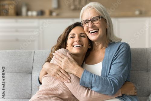 Cheerful attractive senior mom and happy adult daughter hugging at home with love, care, resting on couch together, smiling, laughing, talking, enjoying family relationship, bonding, motherhood photo