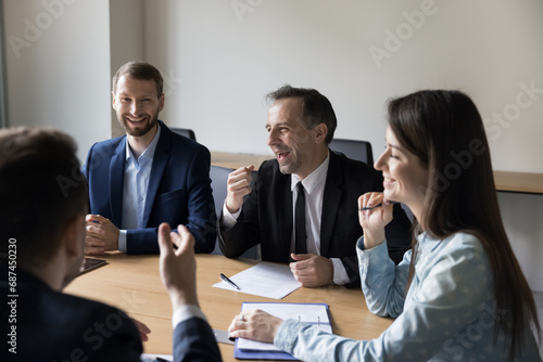 Group of businesspeople laugh at funny joke in boardroom, talk, take break at briefing, enjoy work moments with friendly colleagues, at corporate seminar. Teamwork, companionship teambuilding event