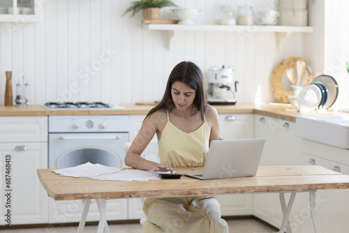 Young serious woman sit at table in kitchen do accounting work, calculates finances, use calculator, fills data result use laptop application, manage family budget, control personal savings, pay bills