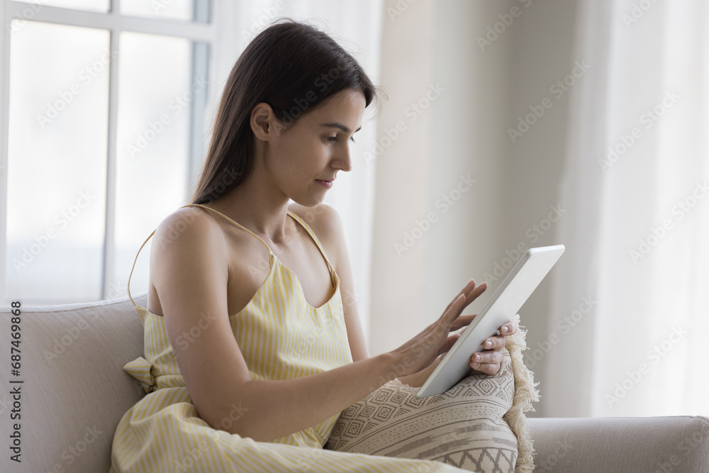 Focused single woman using digital tablet seated on sofa at home, enjoy e-shopping services, booking hotel, planning vacation, buy tickets, teleworking using internet and modern device for online work Focused single woman using digital tablet seated on sofa at home, enjoy e-shopping services, booking hotel, planning vacation, buy tickets, teleworking using internet and modern device for online work